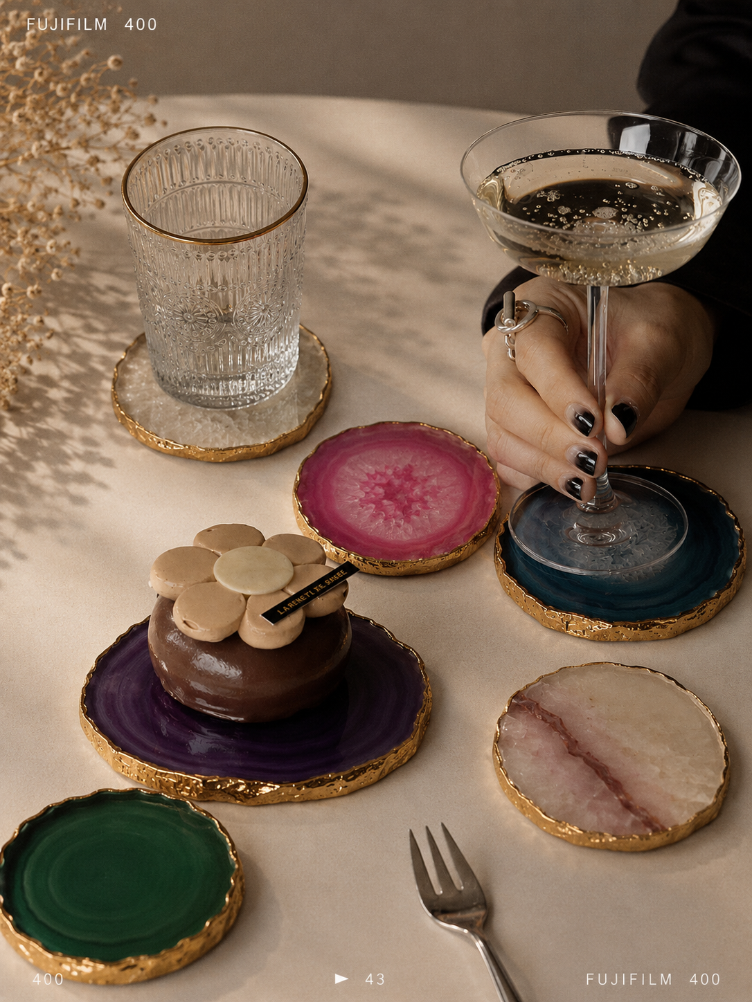 A set of round agate stone coaster plates with gold glitter edges, displayed on a beige tablecloth with lifestyle props including a glass, a coupe glass, a small chocolate dessert, and a fork. A hand with black nail polish is holding the blue coaster under the coupe glass.