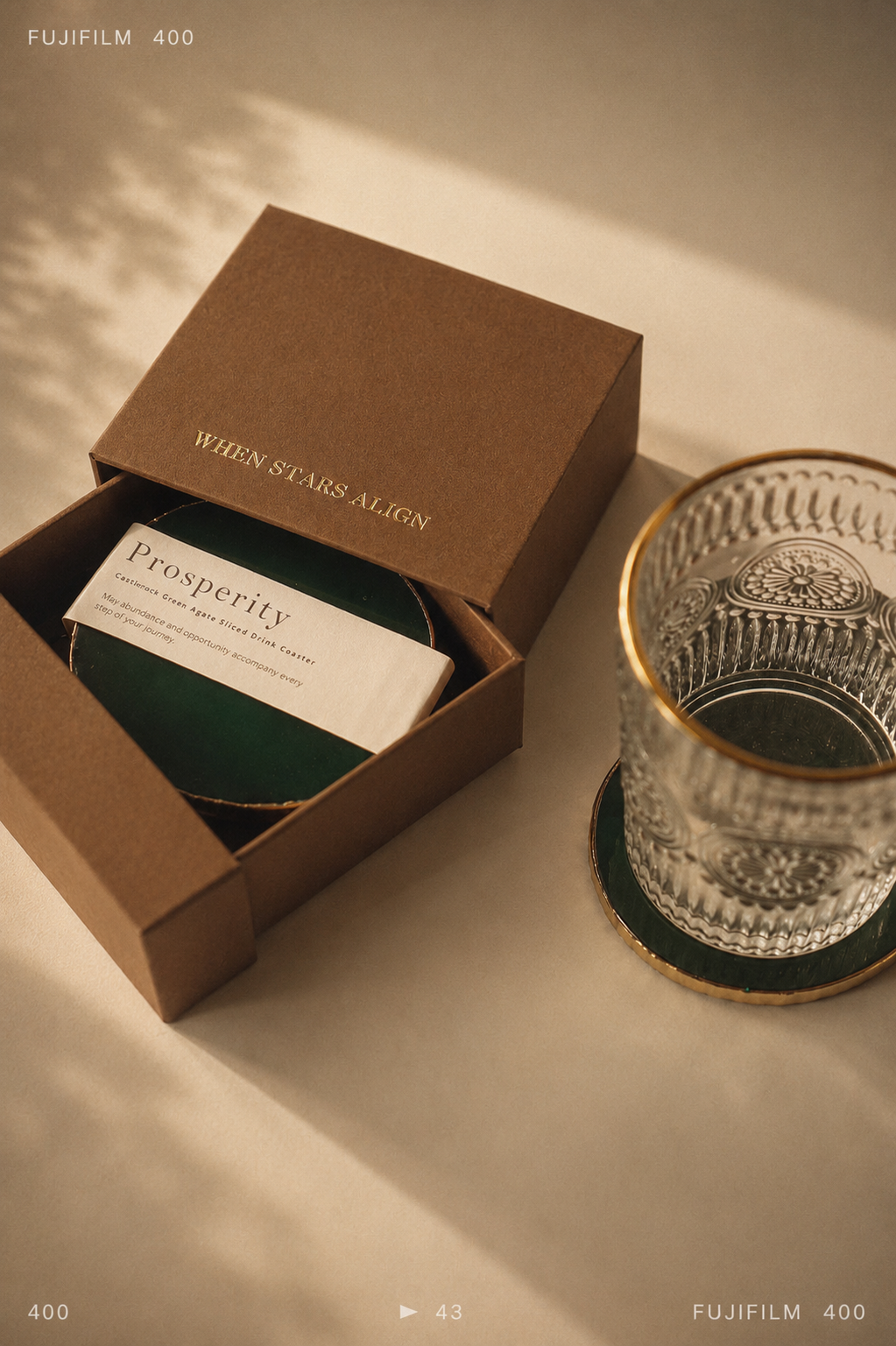A brown kraft gift box labeled 'WHEN STARS ALIGN' is open to reveal a dark green coaster and a white card reading 'Prosperity.' A clear cut-glass tumbler with a gold rim sits beside it on a matching coaster against a warm beige background.